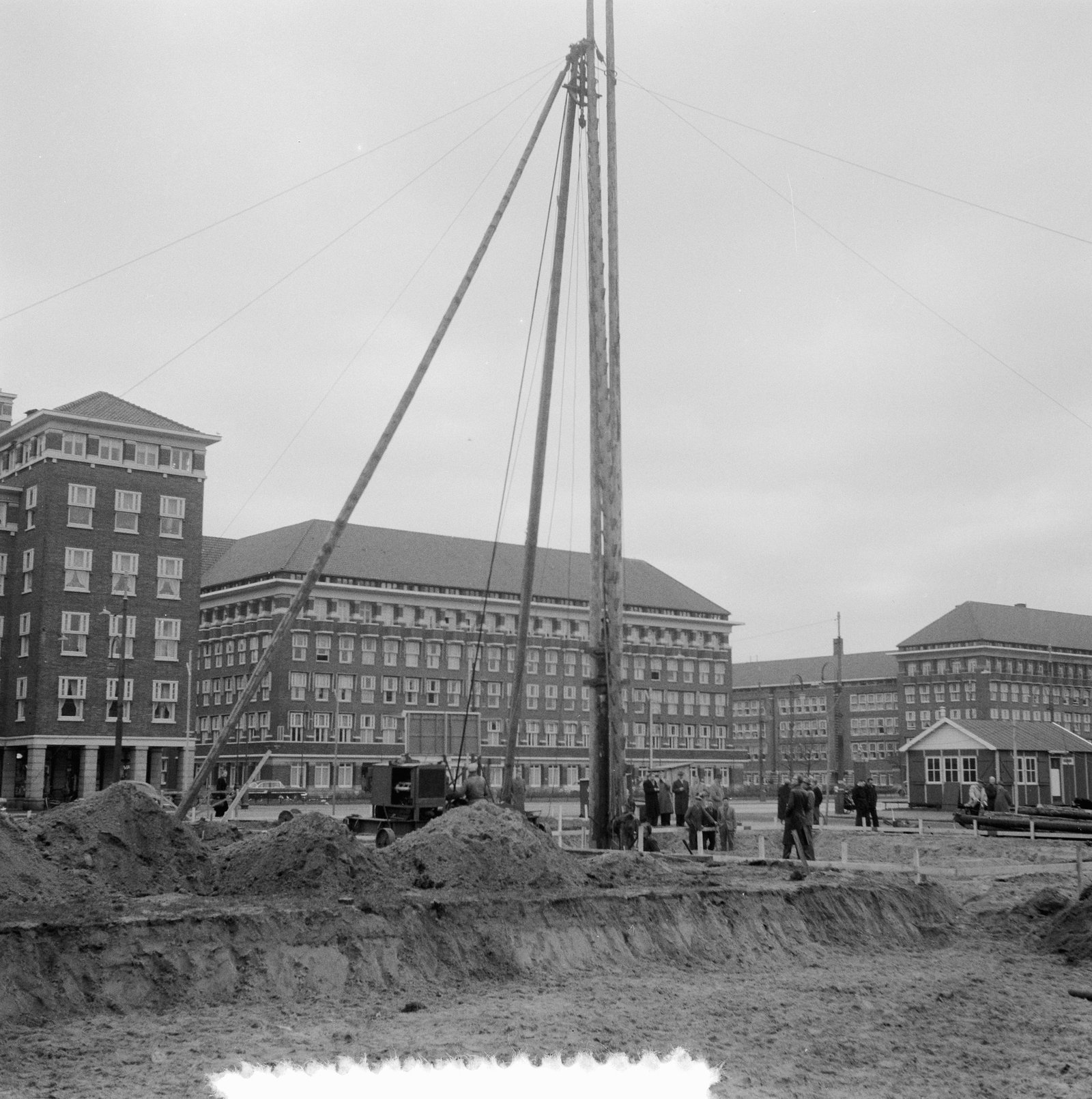 Black-and-white photo of a pile driver on a sandy site, with the pre-war apartment blocks behind it